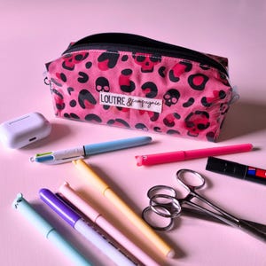May include: Pink pencil case with a black zipper and a skull and leopard print design. The pencil case is next to a white AirPod case, pens, scissors, and markers. The text "LOUTRE & compagnie" is on the pencil case.