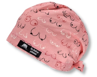 May include: Pink surgical scrub hat with a pattern of white and black breasts. The hat has a black label with the text "la loutre piqueuse" in white.