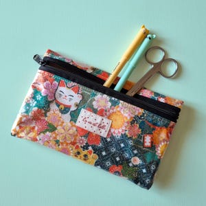 May include: A rectangular pencil case with a black zipper, filled with pens and scissors. The case features a colorful floral pattern with a lucky cat design and the text "La vie est belle."