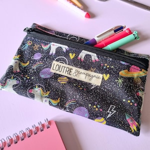 May include: A black pencil case with a zipper, featuring a space-themed pattern with unicorns, planets, and rockets. The text "LOUTRE & compagnie" is visible on a rectangular label. Several pens are partially visible inside the case.