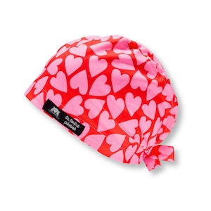 Peut inclure: Un bonnet rouge orné de cœurs roses. Le bonnet est noué dans le dos et porte une étiquette noire avec le texte blanc "la couture piqueuse".