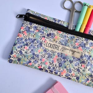 May include: A floral print pencil case with a black zipper, containing scissors and colorful pens. The case features a pattern of blue, pink, and green flowers on a light background. The text "LOUTRE & compagnie" is visible on a label.