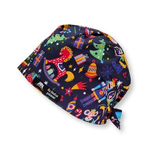 Op de afbeelding: Een marineblauwe operatiemuts met een kleurrijk kerstthema. Het ontwerp bevat een rood schommelpaard, kerstbomen, cadeaus en een notenkraker. De muts heeft een strik aan de achterkant en de tekst "la loutre piqueuse".