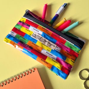 May include: A colorful pencil case with a black zipper, filled with pens and markers. The case is patterned with various drug names and measurements. The text "LOUTRE & compagnie" is visible on a label.