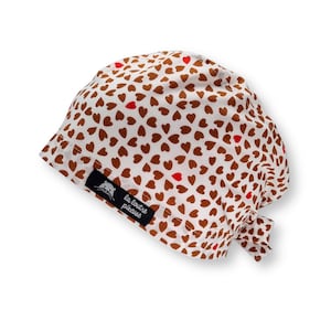 Può includere: Un copricapo bianco con un motivo a cuori marroni e rossi. Un'etichetta nera con un logo di orso e la scritta "la couture piqueuse" è cucita sul lato. Il copricapo è legato sul retro.