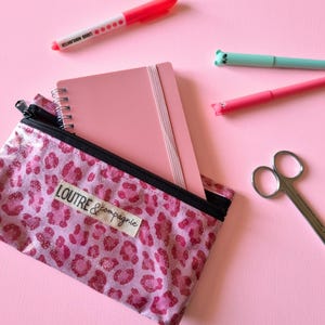 May include: Pink stationery set on a pink background. Includes a pink leopard print pencil case with the text "LOUTRE & compagnie", a pink notebook, pink and teal pens, a pink highlighter, and small scissors.