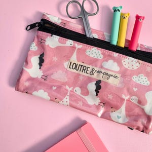 May include: A pink pencil case with a black zipper, featuring a pattern of white storks, clouds, and stars. The text "LOUTRE & compagnie" is visible. Inside are three colorful pens and a pair of scissors.