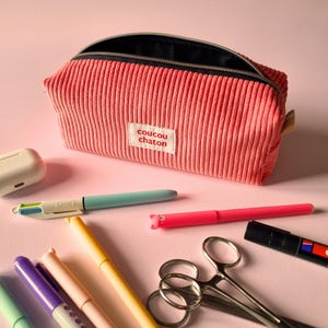 May include: A pink corduroy pencil case with a black zipper, open to reveal the interior. The label reads "coucou chaton". Assorted pens, scissors, and a white earbud case are scattered around the case on a pink surface.
