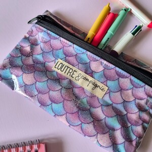 May include: A clear pencil case with a mermaid scale pattern in shades of purple, blue, and iridescent glitter. The case has a black zipper and contains several colorful pens. The text "LOUTRE & compagnie" is on a fabric label.