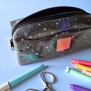 May include: A clear pencil case with a black zipper and a starry night design. The case has a pink label with the text "ZUT, Alors". Assorted pens and scissors are scattered around the case on a light blue surface.