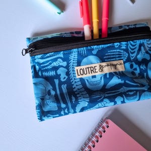 May include: A blue pencil case with a black zipper, featuring a repeating pattern of white skeletons. The pencil case contains several colorful pens. The text "LOUTRE & compagnie" is printed on a small label.