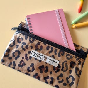 May include: A clear pencil case with a leopard print design, a black zipper, and the text "LOUTRE & compagnie". Inside is a pink spiral notebook with an elastic band. Colorful pens are visible in the background.
