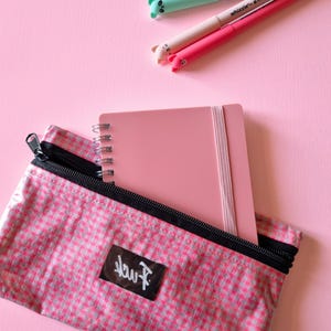 May include: A pink spiral-bound notebook with an elastic closure is partially inside a pink and white checkered pencil case. Three pens with animal-shaped tops are on a pink background. The pencil case has a black zipper and a black label with white text.