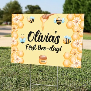 May include: A yellow yard sign with the text "Olivia's First Bee-day!" features illustrations of bees, honey, and honeycomb. The sign is held up by a metal stake and is placed on a grassy lawn.