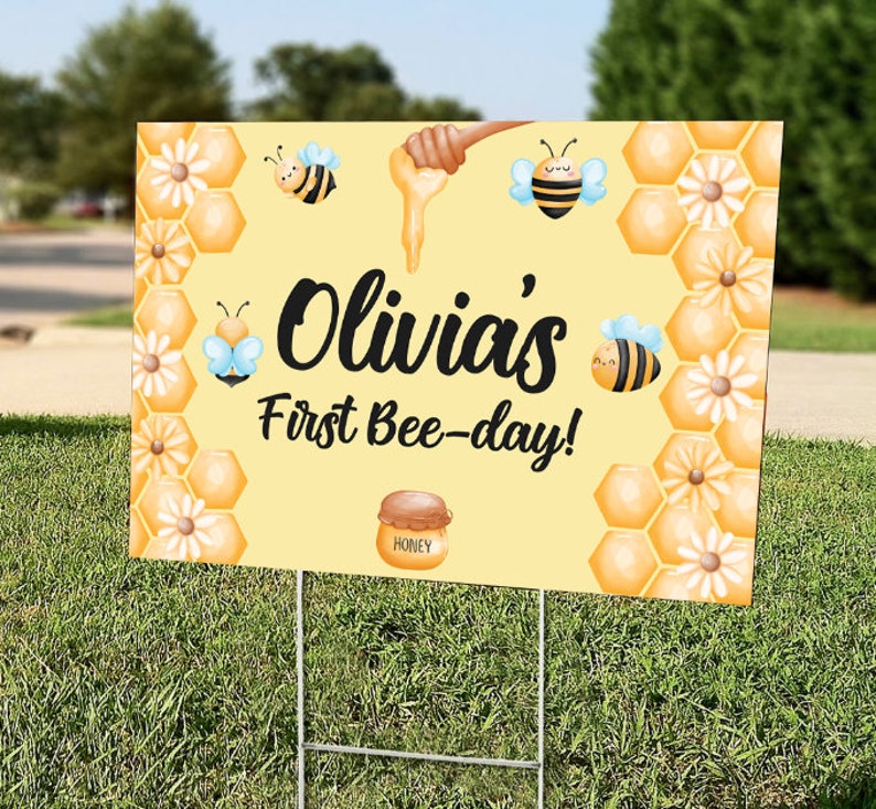 May include: A yellow yard sign with the text "Olivia's First Bee-day!" features illustrations of bees, honey, and honeycomb. The sign is held up by a metal stake and is placed on a grassy lawn.