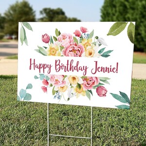 May include: A white sign with a floral design and the text "Happy Birthday Jennie!" printed in burgundy. The sign is on a green lawn.