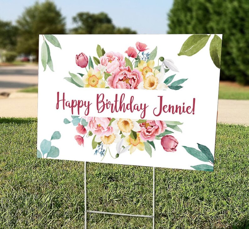 May include: A white sign with a floral design and the text "Happy Birthday Jennie!" printed in burgundy. The sign is on a green lawn.