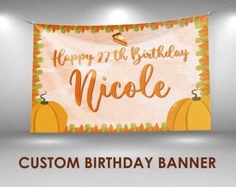 May include: Custom birthday banner with a watercolor background. The banner reads "Happy 27th Birthday Nicole" in orange script. It features orange pumpkins and a slice of pie graphic. The banner is suspended by a wire.