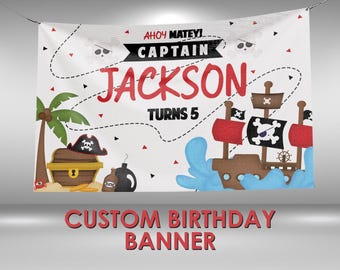 May include: A white pirate-themed birthday banner with red, black, and blue accents. The banner features the text "Ahoy Matey! Captain Jackson Turns 5" and illustrations of a pirate ship, a treasure chest, a palm tree, and a pirate hat.