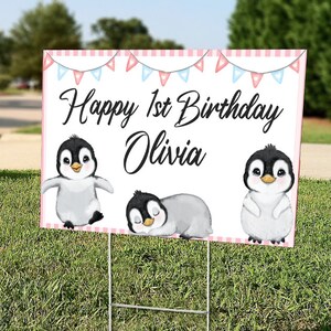 May include: A pink and white birthday sign with a banner of pink and blue flags. The sign says "Happy 1st Birthday Olivia" and features three cartoon penguins.