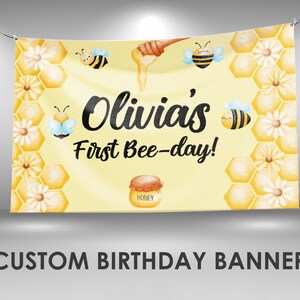 May include: A yellow banner with the text "Olivia's First Bee-day!" features illustrations of bees, honeycombs, and flowers. A honey pot graphic is at the bottom. The banner is suspended by a wire.