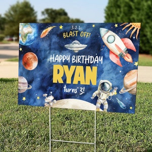 May include: A blue and yellow birthday sign with a space theme. The sign says "Happy Birthday Ryan turns 3!" with a rocket, astronaut, planets, and stars. 3-2-1- Blast Off! is written at the top.