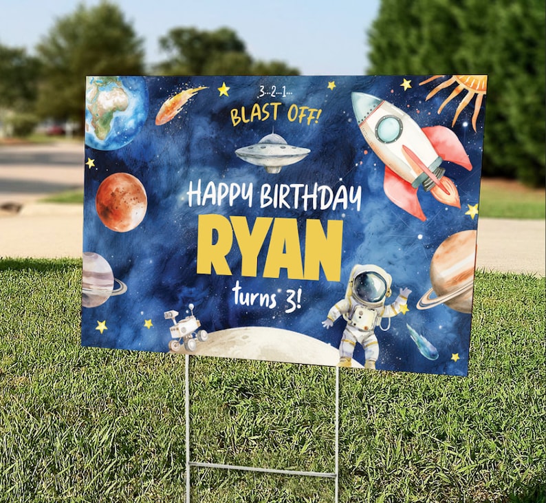 May include: A blue and yellow birthday sign with a space theme. The sign says "Happy Birthday Ryan turns 3!" with a rocket, astronaut, planets, and stars. 3-2-1- Blast Off! is written at the top.