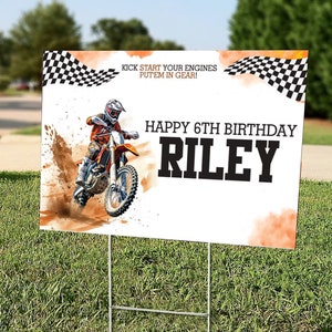 May include: A white and orange birthday sign with a checkered flag design. The sign features a photo of a dirt bike rider and the text "Kick start your engines put'em in gear! Happy 6th Birthday Riley".