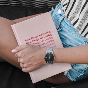 May include: A pink journal with the text "Good Things are Coming" repeated in a stacked pattern. The journal is held by a person wearing a striped shirt and denim shorts. A silver watch with a black face is on the person's wrist.