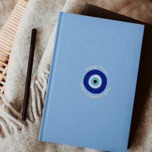 Op de afbeelding: Een lichtblauw notitieboek met een blauw en wit boze oogontwerp en de tekst "energy flows where intention goes".