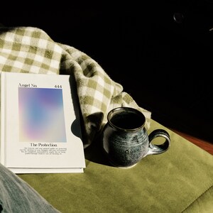 May include: A white book with a holographic cover titled "Angel No. 444 The Protection". The book is lying on a green and white checkered blanket and a green couch. A dark blue ceramic mug is sitting on the couch.