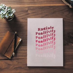 May include: A white notebook with the text "Radiate Positivity" repeated in a gradient of pink and red.