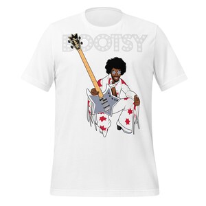 May include: White t-shirt featuring a cartoon illustration of a musician in a white jumpsuit with red star accents, holding a gray bass guitar. The word "BOOTSY" is printed above the illustration in gray.