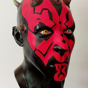 May include: A red and black painted bust of Darth Maul from Star Wars. The bust has horns and yellow eyes.