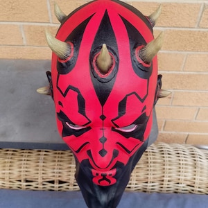 May include: A red and black Darth Maul mask with horns. The mask is made of plastic and has a detailed design.