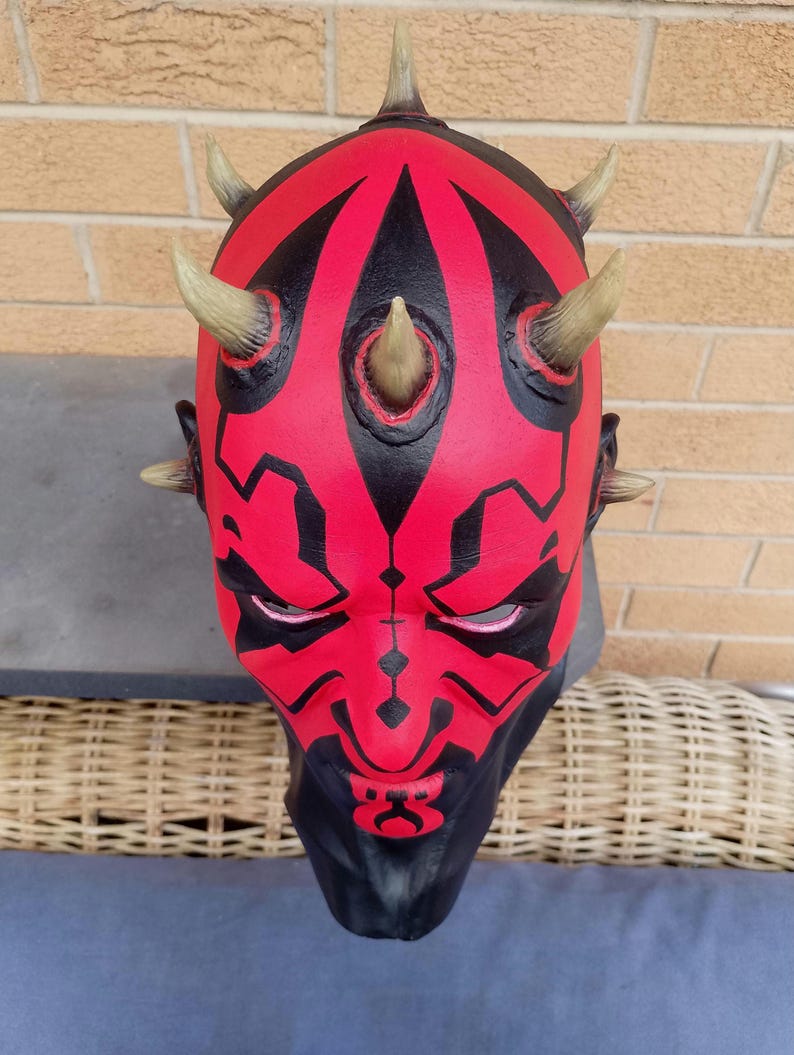 May include: A red and black Darth Maul mask with horns. The mask is made of plastic and has a detailed design.