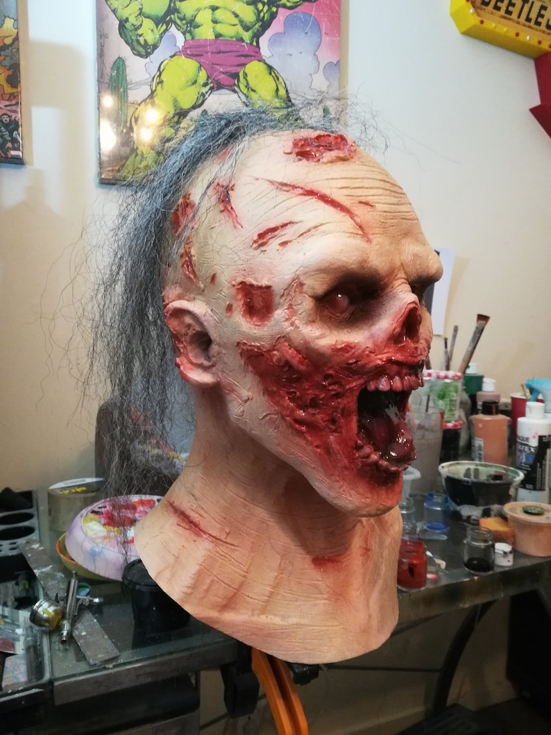 May include: A detailed, realistic zombie mask with a pale, flesh-toned face, red and bloody wounds, and a gaping mouth with a visible tongue. The mask has long, gray hair.