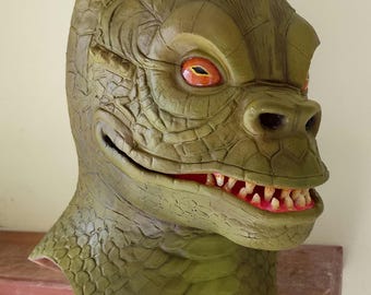 May include: Green latex monster mask with a toothy grin and orange eyes. The mask has a textured surface that resembles scales.