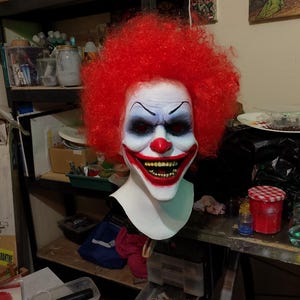 May include: A clown mask with a red curly wig. The mask features a white face with blue eyeshadow, a red nose, and a wide, menacing grin showing yellow teeth. The mask is designed to look scary.
