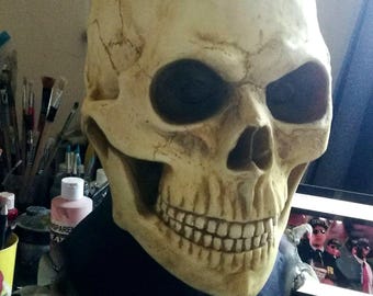 May include: A detailed, off-white skull mask with dark eye sockets and a full set of teeth. The mask is worn over a dark blue garment. The skull has visible cracks and imperfections, giving it a realistic appearance.