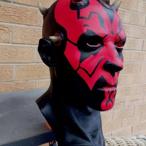 May include: A red and black Darth Maul mask with horns. The mask is made of plastic and has a detailed paint job.