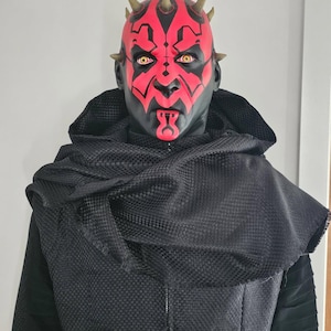 May include: A red and black mask with horns and yellow eyes. The mask is worn by a person with a black hooded robe.