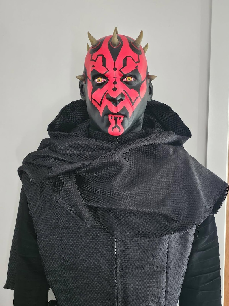 May include: A red and black mask with horns and yellow eyes. The mask is worn by a person with a black hooded robe.