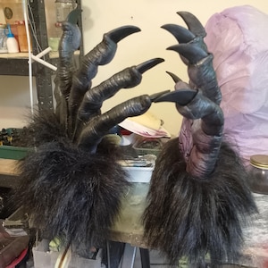 Puede incluir: Par de manos de monstruo negras con garras largas y curvas. Las manos tienen piel texturizada y una base oscura y peluda. Las garras son de color negro brillante y las manos parecen estar hechas de un material duradero.