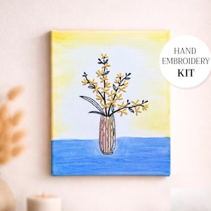Könnte beinhalten: Handstick-Set mit einer Leinwand mit floralem Design. Das Kunstwerk zeigt eine Vase mit gelben Blumen vor einem gelb-blauen Hintergrund. Der Text "HAND EMBROIDERY KIT" befindet sich in einem weißen Kreis.