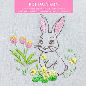 Könnte beinhalten: Stickmuster eines weißen Kaninchens mit rosa Ohren, das in einem Feld mit gelben Blumen sitzt. Das Kaninchen ist von grünen Blättern und rosa Blumen umgeben. Das Muster eignet sich für Stickprojekte.