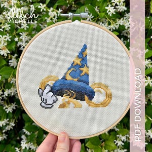 Könnte beinhalten: Kreuzstich-Stickerei in einem Holzrahmen mit einem blauen Zaubererhut mit goldenen Sternen und einer weißen behandschuhten Hand. Der Text "stitch MICHELLE" befindet sich oben links. Die Worte "PDF DOWNLOAD" stehen auf der rechten Seite.