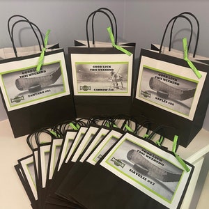 Peut inclure: Un ensemble de 10 sacs cadeaux noirs et verts avec des étiquettes blanches. Les étiquettes présentent un design de bâton de hockey et de rondelle et le texte "Bonne chance ce week-end". Les sacs sont décorés d'un ruban vert.