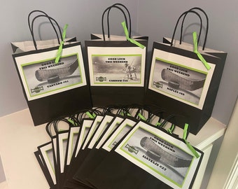 May include: A set of 10 black and green gift bags with white labels. The labels have a hockey stick and puck design and the text "Good Luck This Weekend". The bags are decorated with green ribbon.