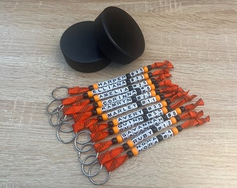 May include: A set of 14 personalized hockey keychain bracelets with orange and black ribbon, white beads, and black letter beads spelling out names and numbers.